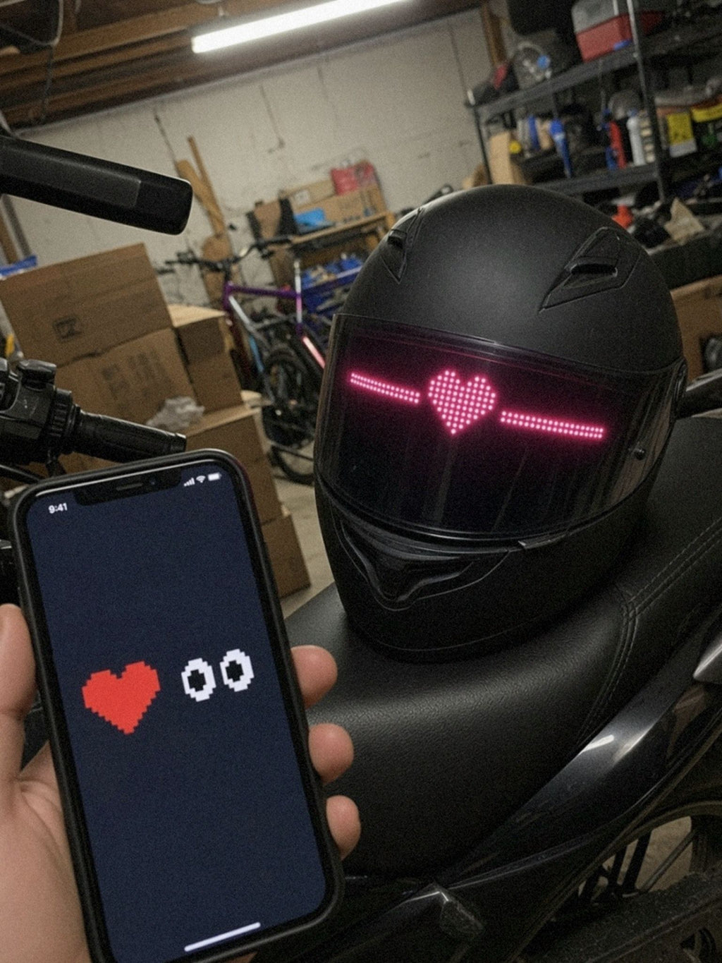 Telefoon met de app in de hand, op de achtergrond een motorhelm op het zadel van een geparkeerde motor met een pixelhartje op het LED-vizierscherm