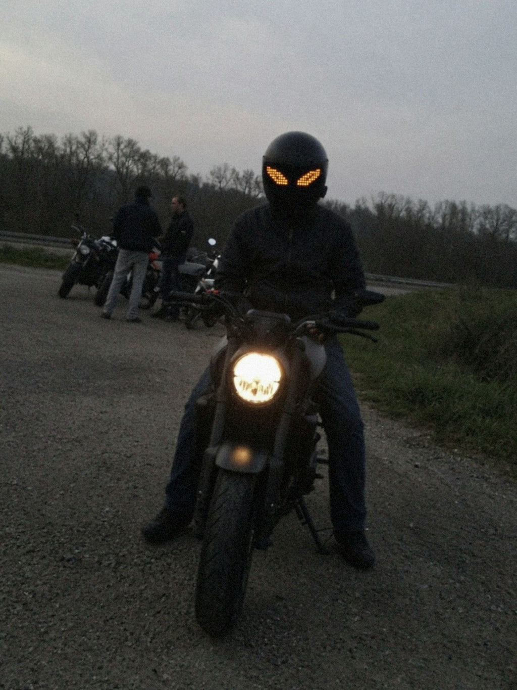 Motorrijder zit met beide voeten aan de grond op zijn stilstaande motor bij een stopplaats in de schemering, het LED-vizierscherm toont oranje pixelogen