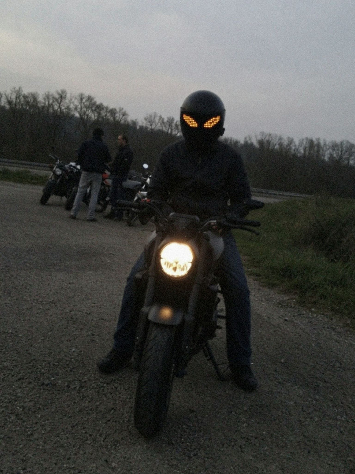 Motorrijder zit met beide voeten aan de grond op zijn stilstaande motor bij een stopplaats in de schemering, het LED-vizierscherm toont oranje pixelogen