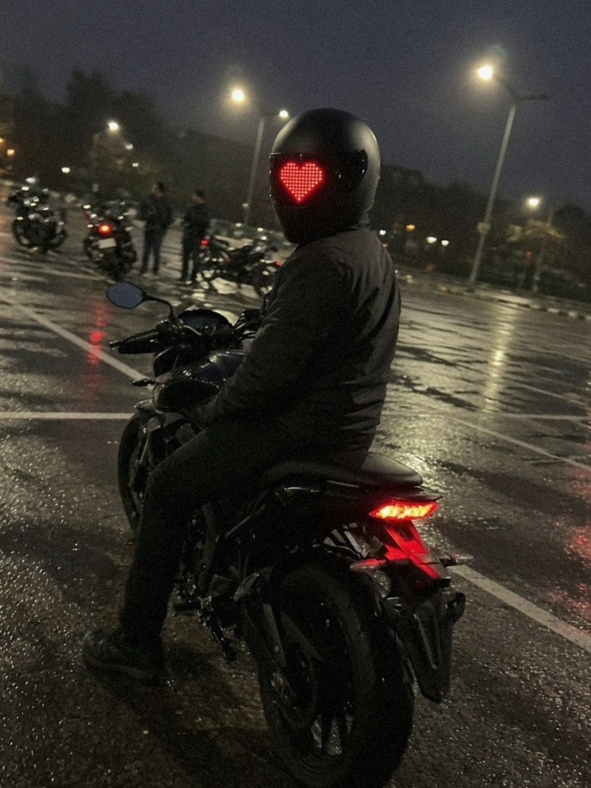 Van achteren gefotografeerd: motorrijder op zijn geparkeerde motor op een natte parkeerplaats kijkt om, het LED-vizierscherm toont een rood pixelhart