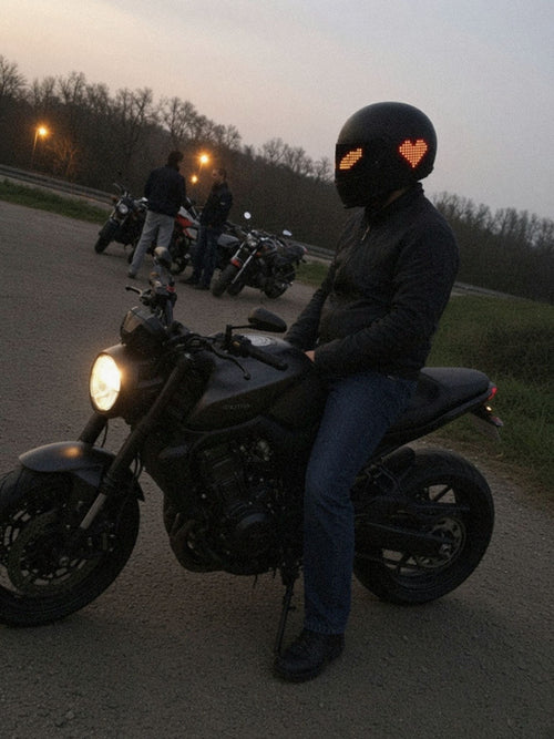 Motorrijder zit op zijn stilstaande motor bij een stopplaats, op zijn helm de LED-vizierstrip met oranje pixelogen en het LED-achterpaneel met een pixelhart